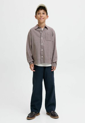 Ragazzo in piedi che indossa un cappello beige, una camicia a quadri con bottoni, pantaloni larghi scuri e scarpe marroni, sorridendo leggermente, su uno sfondo bianco uniforme.