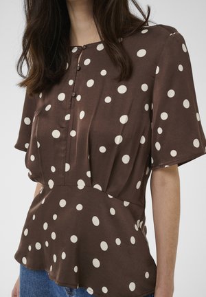 Blouse marron à manches courtes avec des pois blancs, col boutonné et taille légèrement évasée, portée avec un jean bleu.