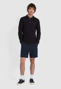 Zwarte poloshirt met lange mouwen, voorzien van een twee-knopen sluiting en klein logo; gecombineerd met gestructureerde marineblauwe shorts en marineblauwe sneakers met witte zolen.