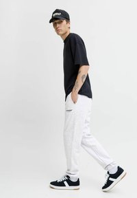 Crni oversize majica, svijetlosive trenirke s brendom i crne tenisice s bijelim akcentima. Model nosi crnu kapu.