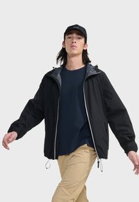 Veste légère noire avec une doublure intérieure grise, fermeture éclair à l'avant, capuche ajustable et poignets élastiques, portée sur une chemise bleu marine et un pantalon beige.