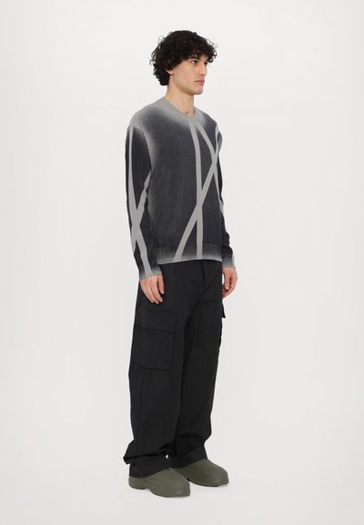 Dunkelgrauer Strickpullover mit hellgrauen abstrakten Streifen, kombiniert mit schwarzen Cargo-Hosen und olivgrünen Schuhen, vor einem weißen Hintergrund.