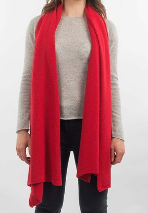 Persona che indossa una maglia a maniche lunghe grigio chiaro, pantaloni neri e una lunga sciarpa rossa drappeggiata intorno al collo su sfondo bianco.