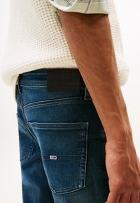 Ciemnoniebieskie dżinsy z gładką fakturą, z czarną etykietą, widocznym przeszyciem i małym logo na tylniej kieszeni.