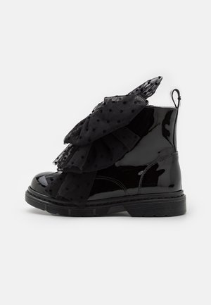 Bottine en cuir verni noir avec des volants en tulle noir à pois superposés sur le devant et une bride d’extraction au talon.
