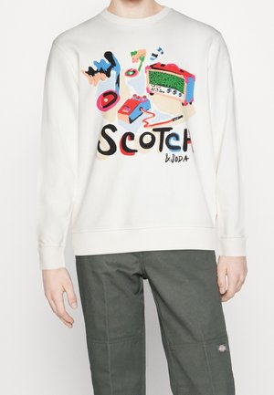Sweat-shirt blanc avec un graphisme abstrait coloré de magnétophones et le texte "Scotch & Soda" porté avec un pantalon vert foncé.