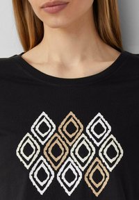 Schwarzes T-Shirt mit einem geometrischen Diamantmuster in Weiß, Creme und Braun, mit Rundhalsausschnitt und weichem Material.