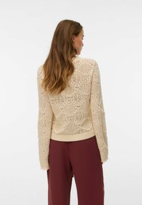 Femme aux longs cheveux bruns portant un pull beige à motifs tricotés et un pantalon ample bordeaux, de dos sur un fond blanc uni.