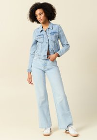 Chaqueta de mezclilla azul claro con mangas largas y dos bolsillos en el pecho, combinada con unos jeans acampanados a juego y zapatillas blancas, sobre un fondo neutro.