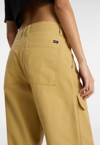 Pantaloni cargo gialli in tela con grandi tasche posteriori, cuciture a vista e un piccolo etichetta Vans vicino alla vita. Design minimalista.