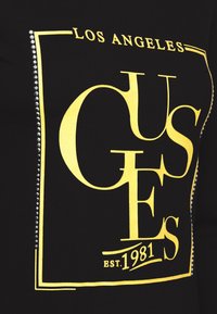 Černá tkanina s žlutým obdélníkovým obrysem a textem „Los Angeles GUESS EST. 1981“, zdobená malými stříbrnými hroty po stranách.