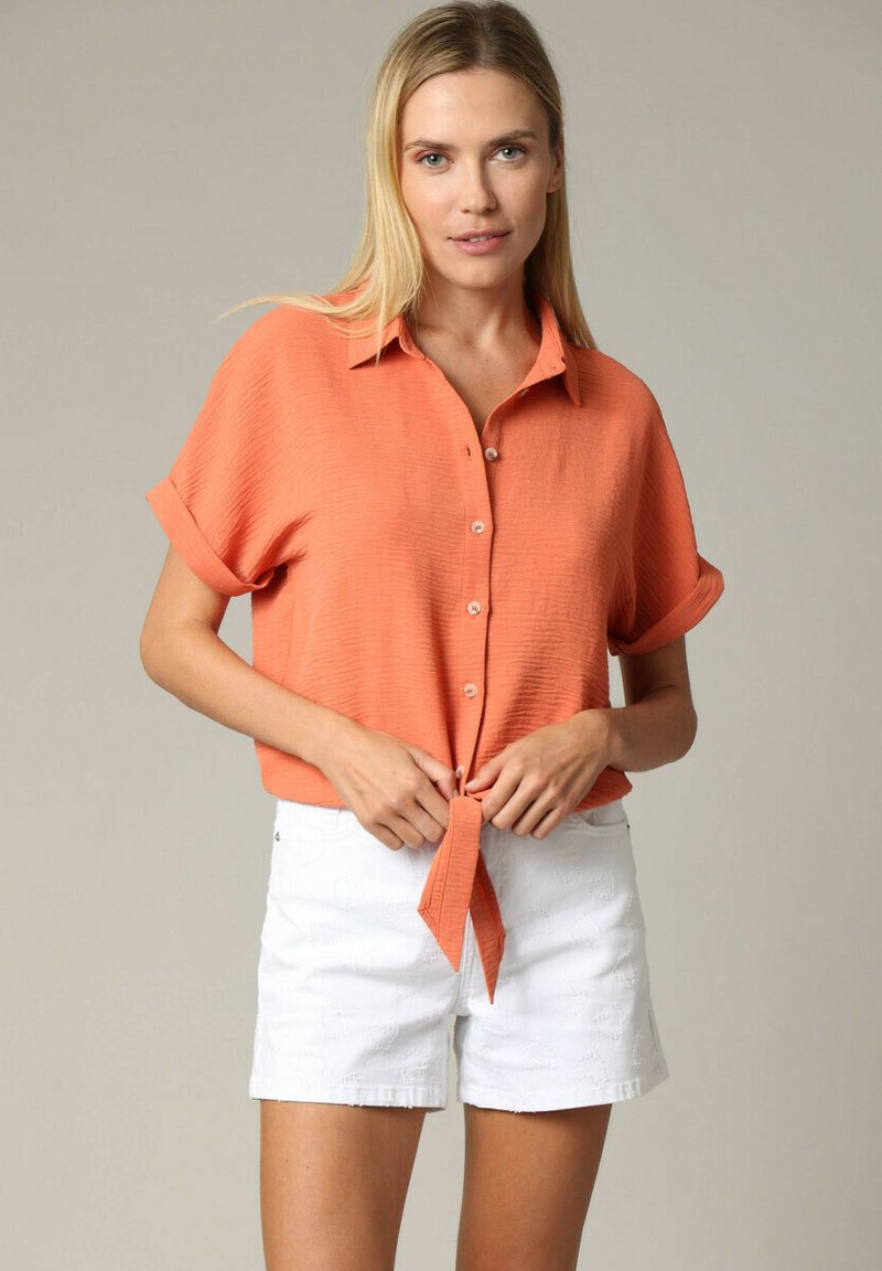 Korte mouwen, button-up shirt in fel oranje, met een gestructureerde stof en een gestrikte zoom, gecombineerd met witte, gestructureerde shorts.