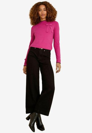 Fuchsia Pullover mit langen Ärmeln und Schleifendetail, kombiniert mit weiten schwarzen Hosen und schwarzen Absatzstiefeln. Flacher, strukturierter Stoff mit gewelltem Rand.