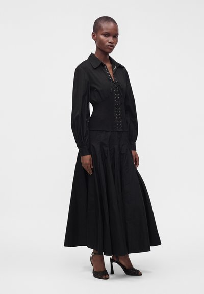 Femme portant un chemisier noir à manches longues avec un détail corset à lacets, une longue jupe noire et des sandales noires à talons hauts.
