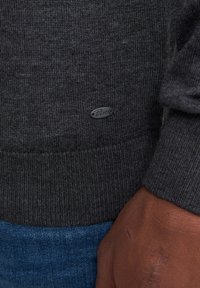 Suéter de punto gris oscuro con dobladillo acanalado, que presenta una pequeña etiqueta con logo plateado. La textura es suave y uniforme.