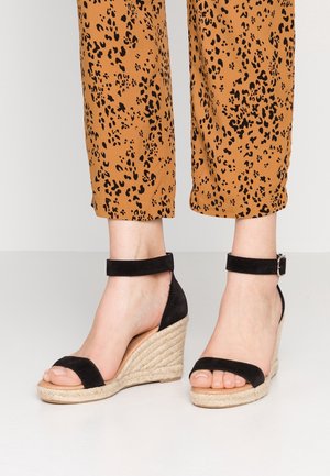 Sandali con zeppa nera in suede, con cinturino alla caviglia e dettagli in corda di juta, caratterizzati da un design con punta aperta. Abbinati a pantaloni cropped con stampa leopardata marrone.