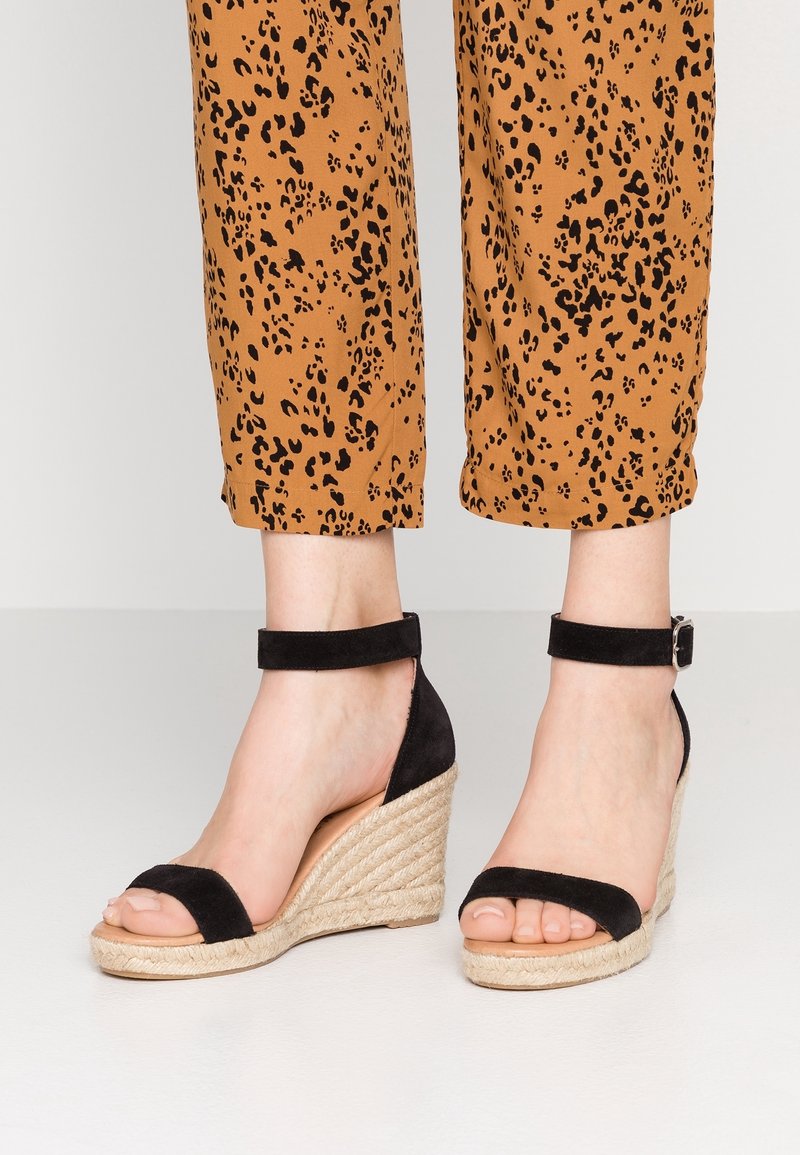 Sandali con zeppa nera in suede, con cinturino alla caviglia e dettagli in corda di juta, caratterizzati da un design con punta aperta. Abbinati a pantaloni cropped con stampa leopardata marrone.