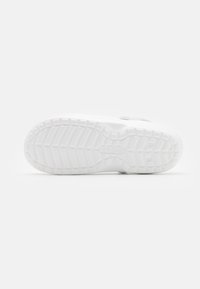 Semelle d'une basket blanche avec motif de grip texturé, détails de la bande de roulement et logo de la marque sur un fond blanc uni.