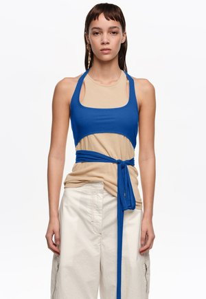 Joven mujer con una camiseta sin mangas beige combinada con un top corto azul anudado y pantalones blancos de pierna ancha, posando frente a un fondo blanco.