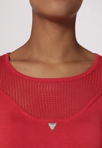 Rode top met korte mouwen, met een textuur en een netachtige halslijn en een klein zilveren driehoeklogo in het midden. Overal gladde stof.