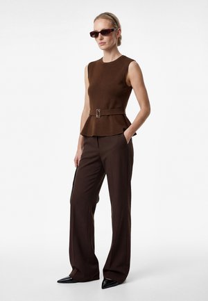 Femme portant un haut marron foncé sans manches ceinturé, un pantalon large assorti, des chaussures noires pointues et des lunettes de soleil foncées, debout les mains dans les poches.