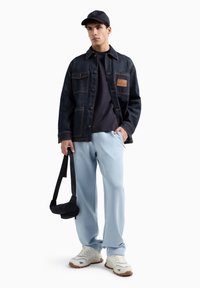Giacca di jeans con tasche sul petto, patch in pelle; felpa blu navy, pantaloni azzurri; cappello nero; sneakers bianche con suola in gomma; borsa a tracolla.