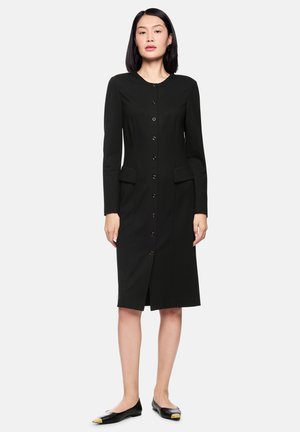 Schwarzes Kleid mit langen Ärmeln, rundem Ausschnitt, vorderen Knöpfen, zwei seitlichen Taschen und knielangem Schnitt. Kombiniert mit schwarzen Ballerinas mit goldenen Details.