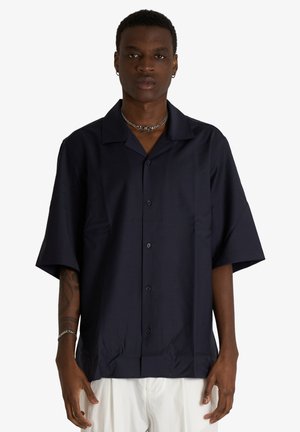 Giovane uomo che indossa una camicia blu navy a maniche corte abbottonata e pantaloni bianchi, in piedi con le braccia rilassate lungo i fianchi su uno sfondo uniforme.