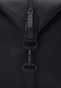 Sac en cuir noir présentant une surface texturée, un design de rabat incliné, une fermeture à glissière en métal et une bandoulière ajustable avec des accents en matériaux métalliques.