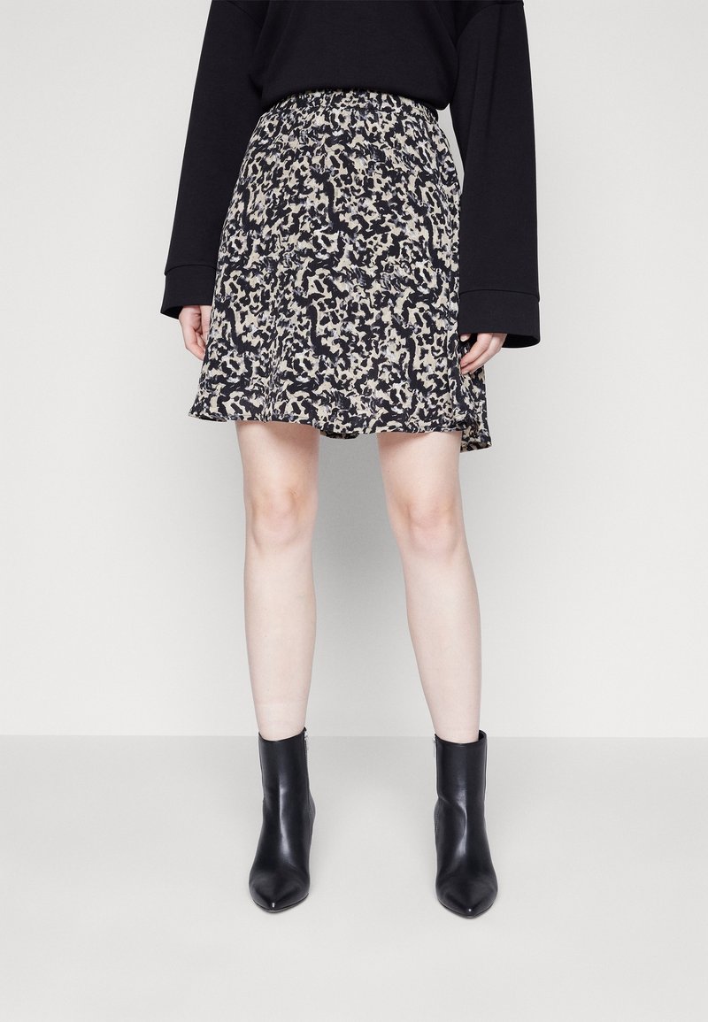 Femme portant une jupe à motifs abstraits noirs et beiges, un haut noir à manches longues, et des bottines noires, debout sur un sol clair uni.