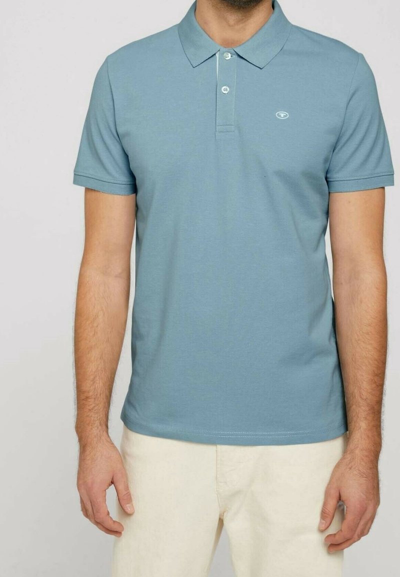 Homme portant un polo bleu clair à manches courtes avec deux boutons et un pantalon beige, debout devant un fond uni.