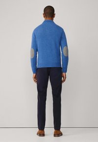 Suéter de punto azul con un cuello alto, que presenta parches de codo grises. Combinado con pantalones oscuros de color marino y zapatos marrones. Diseño simple y casual.