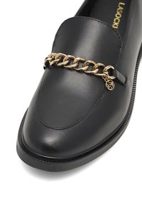 Mocasín de cuero negro con punta redondeada, que presenta un acróstico de cadena dorada en el empeine y un charm decorativo, suela plana.