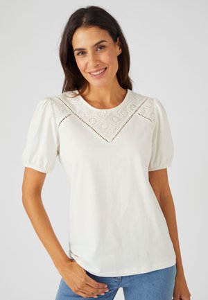 Witte katoenen t-shirt met ballonachtige korte mouwen en een gedetailleerde kantafwerking langs de halslijn, met cirkelvormige patronen en oogjesdetails.