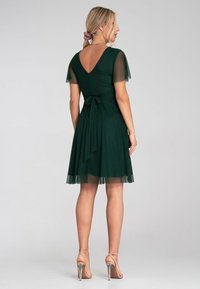 Grünes Kleid mit kurzen, transparenten Ärmeln und einem V-Rücken. Es hat ein eng anliegendes Oberteil und eine Taillenbindung, einen fließenden Rock und einen transparenten unteren Saum.