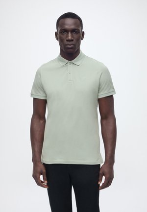 Mężczyzna o ciemnej karnacji, ubrany w jasnooliwkową koszulkę polo z krótkim rękawem i czarne spodnie, stojący na jednolitym białym tle.