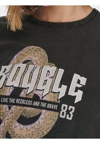 Camiseta negra con un gráfico de serpiente entrelazada con texto audaz y estilizado que dice "TROUBLE" y "83", con una impresión texturizada.