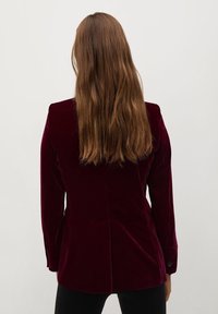 Bordeaux fluwelen blazer met lange mouwen, lichte schouderpadding en een rugventilatie, gecombineerd met zwarte aansluitende broek.