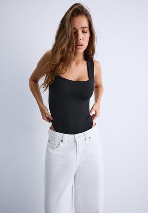 Femme aux cheveux longs ajustant un haut noir sans manches, portant un pantalon blanc sur un fond clair uni.