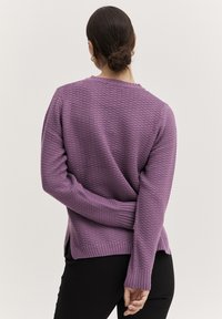 Lila stickad tröja med en texturerad design, ribbade muddar och sidslitsar; bärs över svarta byxor som visar en avslappnad passform.