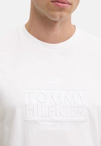 T-shirt bianco in cotone con un logo testurizzato e in rilievo che reca la scritta "TOMMY HILFIGER" e "Dal 1985" al centro. Tessuto liscio con una vestibilità classica.
