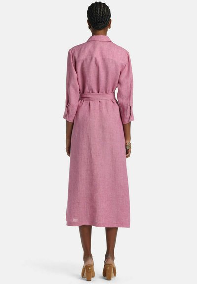 Frau mit geflochtenem Haar, die ein langärmliges, tailliertes rosa Midikleid und braune High-Heels trägt, steht mit dem Rücken zur Kamera vor weißem Hintergrund.