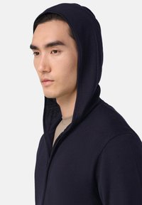 Tamsiai mėlyna zip hoodie su tekstūruotu audiniu, turintis gobtuvą ir pilno ilgio užtrauktuką. Modelis vilki béžinį marškinėlį po juo.