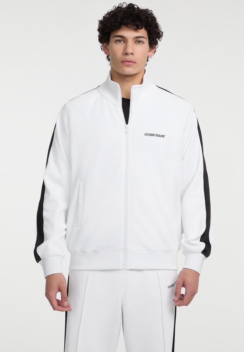 Witte zip-up jas van gladde stof, met zwarte zijkanten, een hoge kraag en geborduurd "GUESS JEANS" logo op de borst.