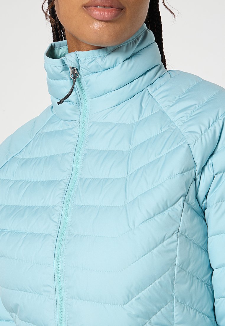 Mujer vistiendo una chaqueta acolchada de color azul claro con un cuello alto con cremallera y mechones de cabello trenzado visibles.