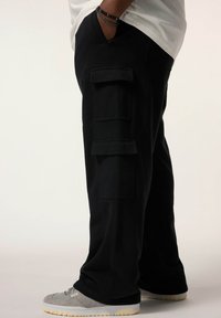 Schwarze Cargo-Hosen mit mehreren Seitentaschen und einer lockeren Passform. Weiches Material und eine glatte Textur. Kombiniert mit grauen Sneakers.