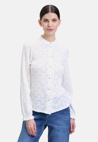Blusa bianca in pizzo con colletto alto, chiusura frontale con bottoni e polsini arricciati, caratterizzata da un intricato motivo a diamante e una texture semitrasparente.