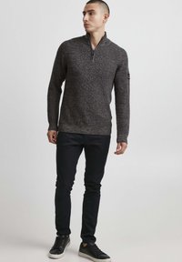 Mørkegrå strikket sweater med lynlås krave, struktureret stof, båret med sorte slim-fit bukser og sorte sneakers. Har et diskret logo på ærmet.