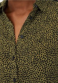 Camisa verde de botones con un patrón moteado en negro, con cuello y botones negros, hecha de una tela texturizada.