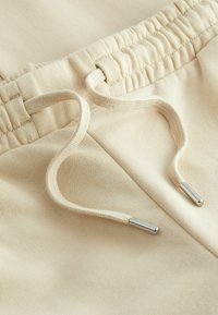 Beige joggingbukser med en tekstureret overflade, der har en elastisk talje og snore med sølvfarvede metalspidser.